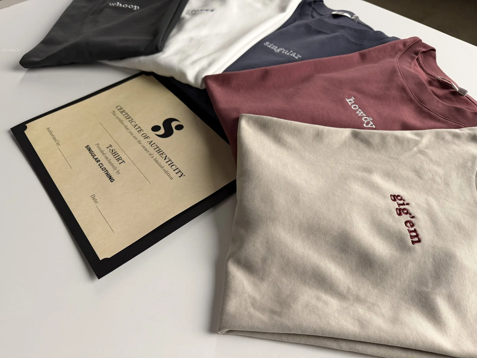Folded embroidered tees and the black certificate folio laid out across a white studio table, seen from an angle