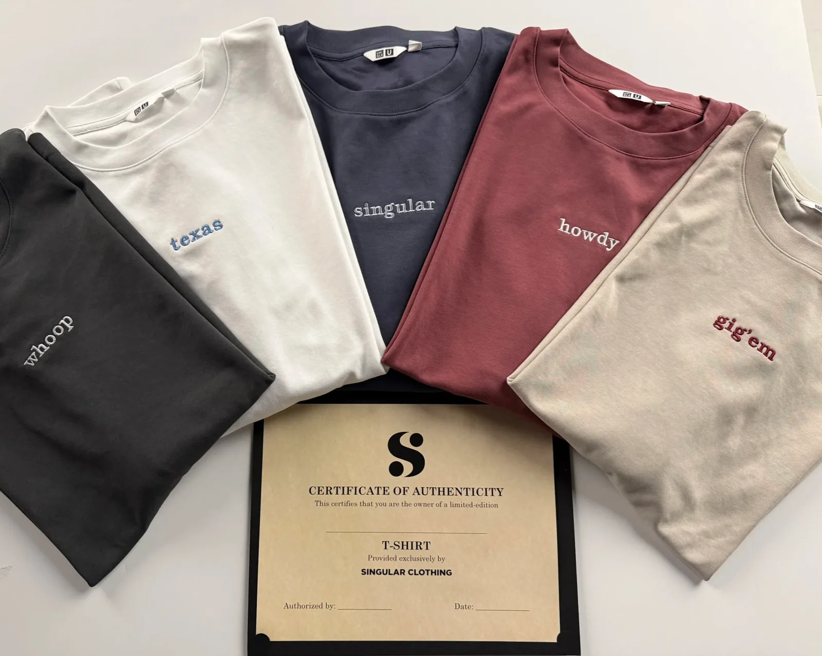 Five folded tees from singular by syntax — whoop in charcoal, texas in white, singular in navy, howdy in brick red and gig 'em in sand — fanned out on the studio table above a certificate of authenticity in its black folio